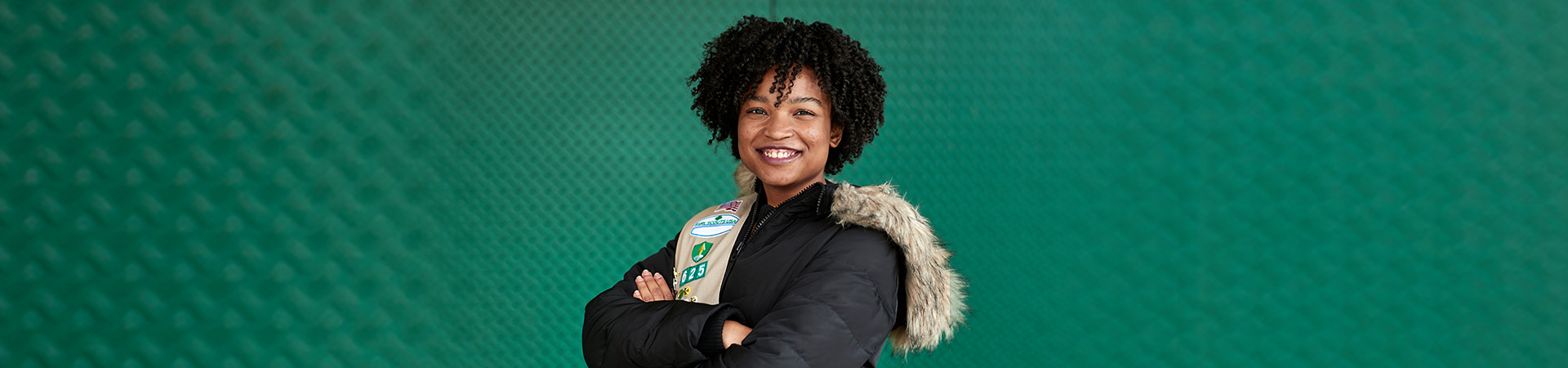  High school girl in Girl Scout sash and winter jacket smiling at camera against green backdrop. 